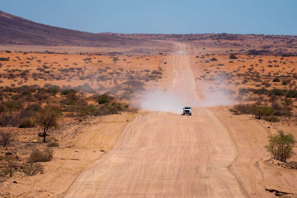 Ein weißer Geländewagen fährt auf einer staubigen Sandstraße durch eine weite, trockene Landschaft unter einem blauen Himmel.