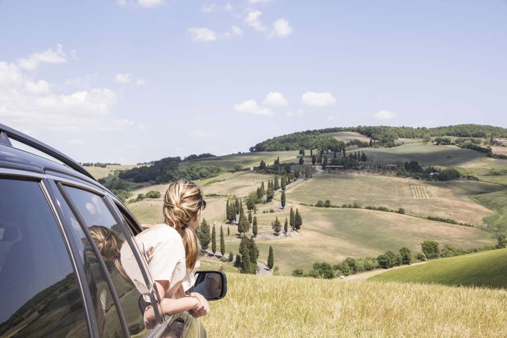 Eine Frau mit blondem Haar lehnt sich aus dem offenen Fenster eines Autos und blickt auf eine hügelige Landschaft mit Zypressen und einem gewundenen Weg.