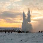 Eine Gruppe von Menschen steht in einem schneebedeckten Feld vor einem Geysir