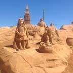 Eine Sandskulptur eines Mädchens und eines Wolfes mit einem großen Uhrturm im Hintergrund