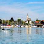 Ein Boot fährt im Vordergrund auf einem ruhigen Gewässer, während im Hintergrund eine Reihe von Segelbooten an einem Steg vertäut ist und eine Stadt mit Kirche und Gebäuden zu sehen ist.