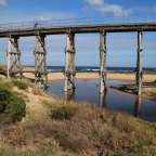 Ein hölzerner Viadukt überspannt einen Flusslauf am Meer unter einem blauen Himmel mit Wolken.