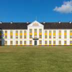 Ein weißes Gebäude mit gelben Akzenten und zahlreichen Fenstern steht vor einem blauen Himmel mit einigen Wolken.