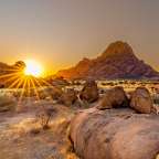 Ein Sonnenuntergang beleuchtet die Felsformationen in einer trockenen Landschaft mit Büschen und trockenem Gras.
