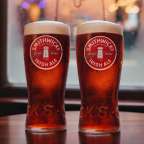 Zwei Gläser Smithwicks Irish Ale stehen auf einem Holztisch vor einem Fenster mit Lichtern im Hintergrund.