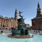 Ein Brunnen auf dem Rathausplatz in Kopenhagen steht vor dem Rathaus und weiteren Gebäuden, während Menschen den Platz beleben.