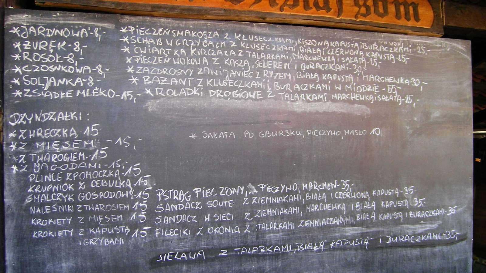 Das Bild zeigt eine Tafel mit einer Speisekarte in weißer Schrift auf schwarzem Grund, die im oberen Bereich durch eine braune Holzverkleidung mit dem Titel "a u nos dzisiaj som" ergänzt wird.