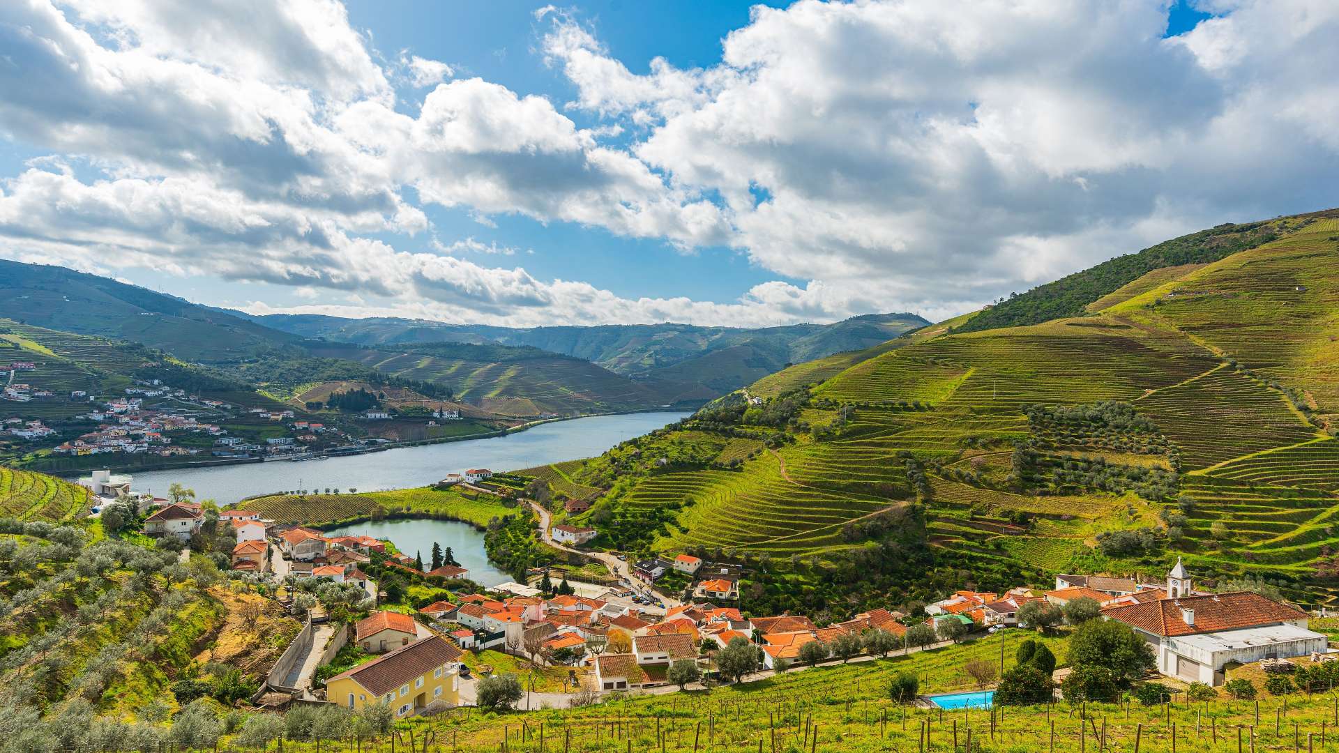 Die malerische Landschaft zeigt ein Dorf mit roten Ziegeldächern, das von terrassierten Weinbergen und einem Fluss umgeben ist, unter einem dramatischen Himmel mit Wolken.