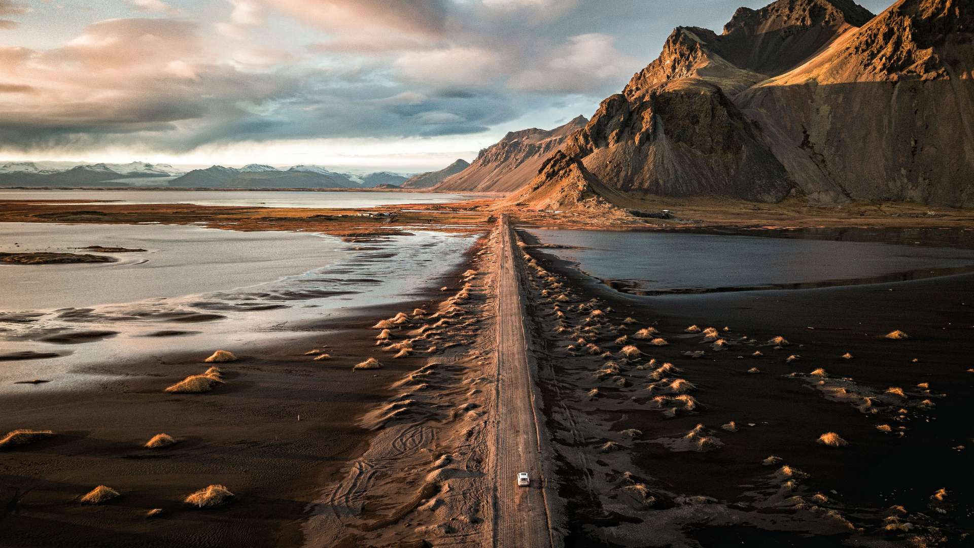 Eine Luftaufnahme zeigt eine Straße, die durch eine schwarze Sandwüste und Gewässer zum Fuße eines schneebedeckten Gebirges führt, das im goldenen Licht der untergehenden Sonne beleuchtet wird.