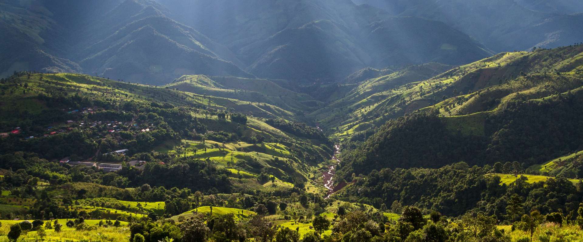 Ein sonnenbeschienener Bergrücken mit einem Dorf und einem Fluss, der sich durch das Tal schlängelt, unter einem dramatisch bewölkten Himmel.