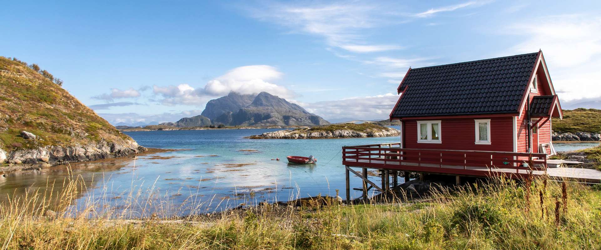 Ein rotes Haus auf Stelzen steht am Ufer eines ruhigen Gewässers mit einem Berg im Hintergrund.