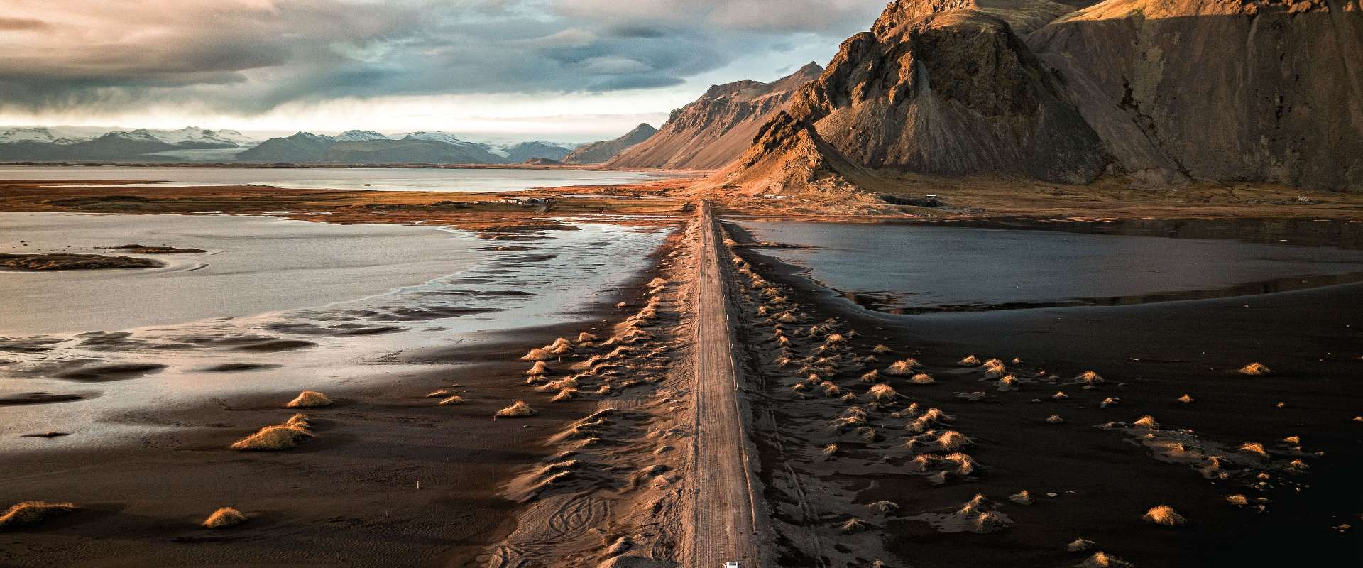 Eine Luftaufnahme zeigt eine Straße, die durch eine schwarze Sandwüste und Gewässer zum Fuße eines schneebedeckten Gebirges führt, das im goldenen Licht der untergehenden Sonne beleuchtet wird.