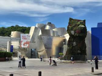 Das Bild zeigt die skulpturale Gestaltung des Guggenheim Museums Bilbao, mit dem Blumen-Welpen als Blickfang und dem blauen Gebäude im Hintergrund.