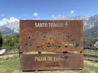 Ein rostfarbenes Schild mit ausgeschnittenen Buchstaben, das "SANTO TORIBIO" und "PICOS DE EUROPA" anzeigt, steht vor einer Bergkulisse und einem blauen Himmel mit Wolken.
