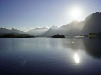 Ein stiller See spiegelt die aufgehende Sonne und die schattigen Umrisse majestätischer Berge in einer ruhigen, friedlichen Landschaft.