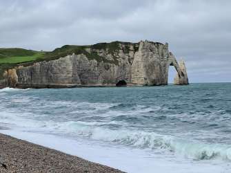 Eine felsige Küstenlandschaft mit einer Bogenfelsenformation im Meer, darunter eine Kiesbucht und das türkisfarbene Meer.