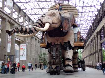 Ein riesiger mechanischer Elefant steht unter einer Stahl- und Glaskonstruktion, umgeben von Besuchern und Plakaten.