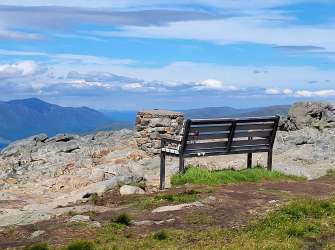 Ein hölzerner Sitzplatz steht auf einem felsigen Berggipfel mit Blick auf einen See und Berge unter einem blauen Himmel mit Wolken.