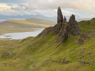 Die dramatischen Felsformationen des Old Man of Storr ragen über eine grüne, hügelige Landschaft mit einem See im Hintergrund auf.
