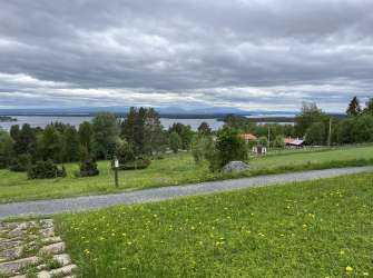 Eine idyllische Landschaft mit einem See, Bergen im Hintergrund und einem grasbewachsenen Hang, auf dem kleine Häuser und Bäume stehen, unter einem dramatischen Himmel mit Wolken.