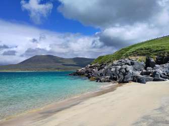 Das Bild zeigt eine felsige Küste mit einem türkisfarbenen Meer, einem sandigen Strand und bewachsenen Hügeln im Hintergrund unter einem bewölkten Himmel.