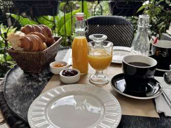 Ein gedeckter Tisch mit Croissants, Orangensaft und Kaffee steht im Freien vor blühenden Blumen.