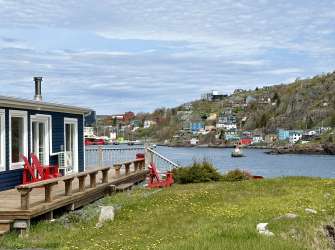 Ein Haus mit einer Veranda und zwei roten Stühlen blickt auf ein Dorf mit bunten Häusern, das an einem steilen Hang liegt, und auf das Meer, auf dem ein kleiner Leuchtturm steht.