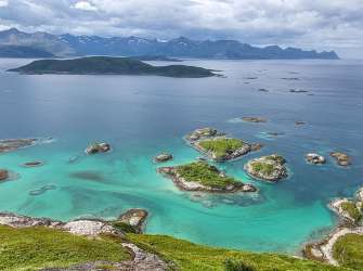 Eine von Inseln übersäte Küstenlandschaft mit türkisfarbenem Wasser und schneebedeckten Bergen im Hintergrund unter einem bewölkten Himmel.
