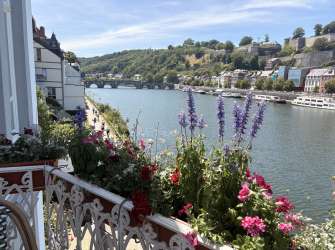 Ein Blick über einen Balkon mit Blumen auf einen Fluss mit Booten, einer Brücke und einer bergigen Stadtkulisse.