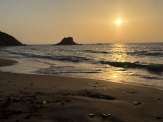 Ein ruhiger Strandblick bei Sonnenuntergang mit Wellen, die auf den Sand treffen, und einem kleinen Felsen im Meer.