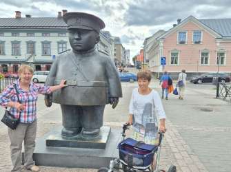 Eine Frau mit einem Rollator steht neben einer Statue eines Mannes in Uniform, während im Hintergrund Gebäude und andere Menschen zu sehen sind.