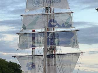 Ein großes Segelschiff mit mehreren weißen Segeln, auf denen blaue Muster und Text zu sehen sind, liegt in einem Kanal, gesäumt von Bäumen und Gebäuden.