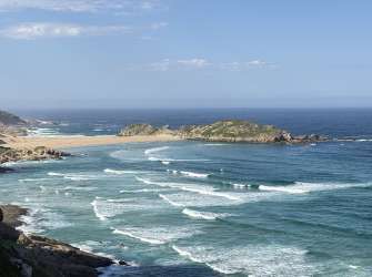 Eine weite Aufnahme einer Küstenlandschaft zeigt schroffe Felsen, einen sandigen Strand und das offene Meer mit sanften Wellen unter einem klaren Himmel.