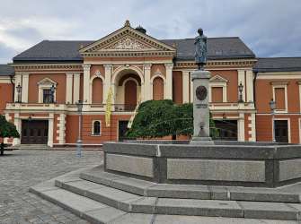 Ein Brunnen und eine Statue stehen auf einem gepflasterten Platz vor einem prächtigen orangefarbenen Gebäude mit einem verzierten Giebel und mehreren Fenstern.