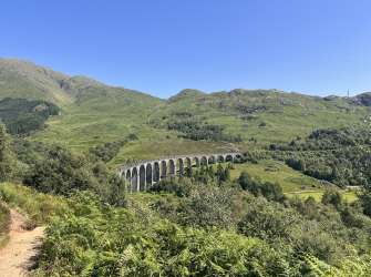 Ein eleganter Eisenbahnviadukt aus Stein mit vielen Bögen erstreckt sich über eine üppig grüne, hügelige Landschaft unter einem strahlend blauen Himmel.