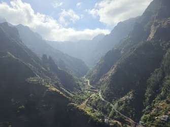 Ein tiefes Tal mit terrassierten Hängen und Häusern erstreckt sich zwischen steilen, bewaldeten Bergen unter einem bewölkten Himmel.