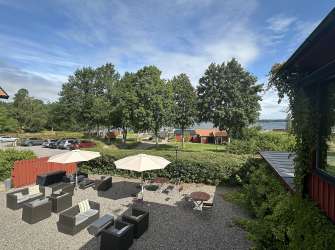 Eine Terrasse mit Sitzgelegenheiten und Sonnenschirmen ist mit Blick auf einen See von Bäumen und roten Holzhäusern umgeben.