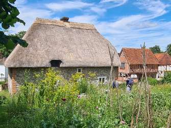 Ein Cottage mit Strohdach steht in einem üppigen Garten vor einer ländlichen Landschaft mit Fachwerkhäusern und einem blauen Himmel mit Wolken.