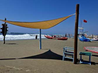 Ein Strand mit einem Sonnensegel, einem Rettungsboot und mehreren Surfbrettern im Sand unter blauem Himmel.
