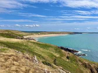 Eine weite Küstenlandschaft mit grasbewachsenen Hügeln, die zum türkisfarbenen Meer abfallen, und einem sandigen Strand mit vereinzelten Personen im Hintergrund unter einem bewölkten blauen Himmel.