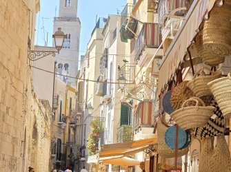 Menschen schlendern am Tag durch eine schmale Gasse in Bari, überragt von einem hohen Kirchturm und belebten Balkonen.