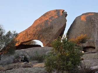 Sonnenbeschienene Felsformationen und ein eingearbeitetes Steingebäude in einer mediterranen Hügellandschaft bei Sonnenuntergang.