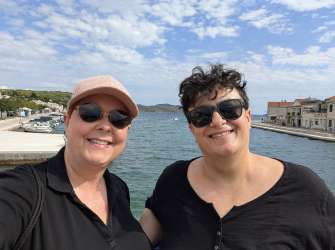 Zwei Personen posieren für ein Selfie auf der Brücke im Küstenort Tisno vor Wasser, Booten und Häusern.