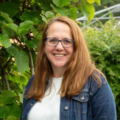 Eine Frau mit Brille und Jeansjacke lächelt vor einem Baum