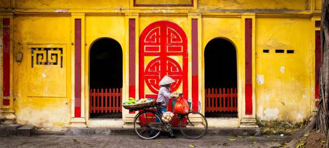 Hanoi: Straßenverkäuferin Eine Frau fährt mit einem Fahrrad vor einem gelben und roten Gebäude