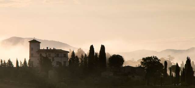 San Cascian, Morgenstimmung: Reisefoto von – ©Birgit und Christopher Eine Silhouette eines Hauses mit Bäumen und Bergen im Hintergrund