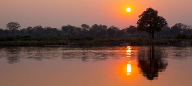 Auwälder, Sümpfe und Flusslandschaften: Caprivi-Streifen Die Sonne geht über einem Fluss mit Bäumen im Hintergrund unter