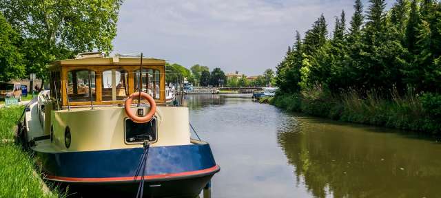 Mit dem Hausboot entdecken: Canal du Midi Ein blau-weißes Boot mit einem weißen Ring am Bug steht an einem Fluss