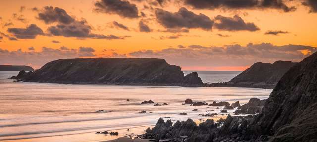 Dramatische Felsküste: Pembrokeshire Coast Ein Sonnenuntergang über einem felsigen Strand mit einem großen Felsen im Hintergrund