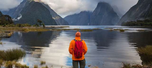 Ein Mann mit einem roten Rucksack steht in der Nähe eines Sees mit Bergen im Hintergrund